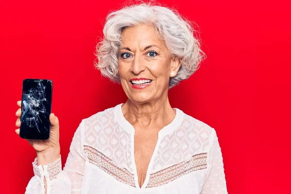 壊れたスマートフォンを持っているシニア白髪の女性は ポジティブで幸せな立っていると歯を示す自信を持って笑顔で笑顔を見せます — ストック写真