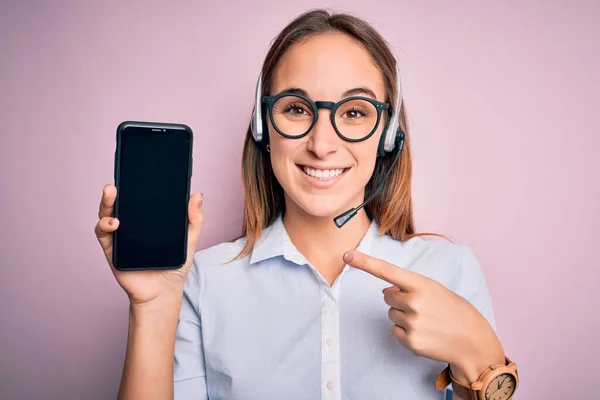 Beautiful call center agent woman working using headset holding smartphone holding screen very happy pointing with hand and finger