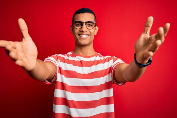 年轻英俊的非洲裔美国男子 身穿休闲条纹T恤和眼镜 微笑着张开双臂拥抱镜头 喜庆的表情拥抱幸福 — 图库照片