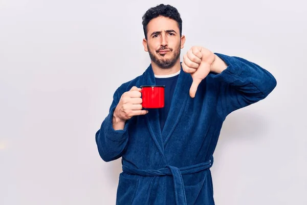 Young hispanic man wearing robe holding coffee with angry face, negative sign showing dislike with thumbs down, rejection concept