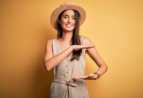 Young beautiful brunette woman on vacation wearing casual dress and hat gesturing with hands showing big and large size sign, measure symbol. Smiling looking at the camera. Measuring concept.