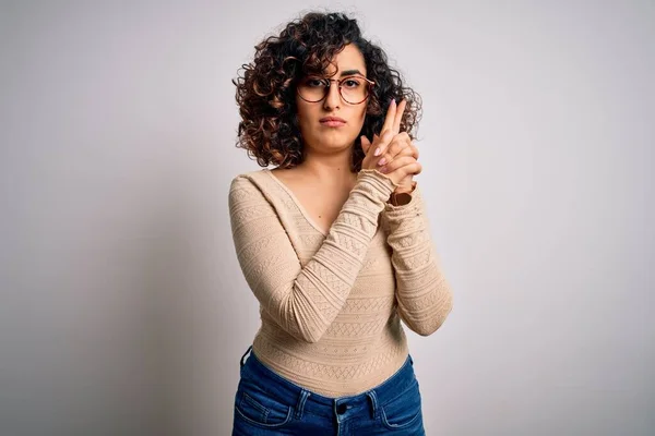 白い背景にカジュアルなTシャツと眼鏡を身に着けている若い美しい巻きアラブの女性手のジェスチャーで象徴的な銃を保持します シューティングゲームをプレイ 怒りの顔 — ストック写真