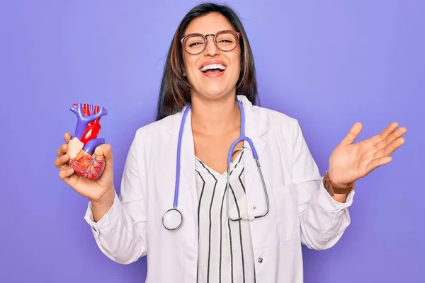 Young doctor cardiology specialist woman holding medical heart over pruple background very happy and excited, winner expression celebrating victory screaming with big smile and raised hands