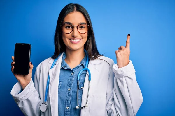 Young beautiful brunette doctor woman wearing glasses and coat holding smartphone surprised with an idea or question pointing finger with happy face, number one