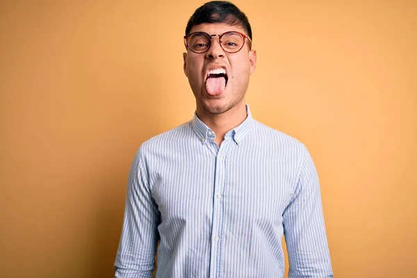 黄色の背景にオタク眼鏡をかけた若いハンサムなヒスパニック系のビジネスマンが面白い表情で幸せ舌を突き出しています 感情概念 — ストック写真