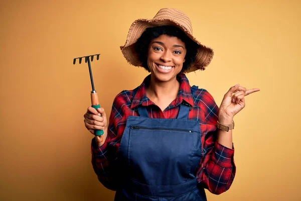 若いですアフリカ系アメリカ人アフロ農家の女性とともに巻き毛を身に着けているエプロンと帽子レーキを使用して非常に幸せなポインティングで手と指で側面 — ストック写真