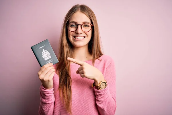 ピンクの背景にカナダのパスポートを保持している美しい赤毛の観光客の女性非常に幸せなポインティングとともに手と指 — ストック写真