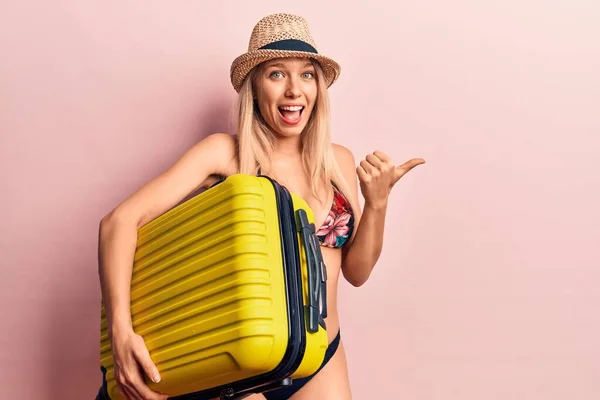 Young beautiful blonde woman wearing bikini and hat holding cabin bag pointing thumb up to the side smiling happy with open mouth