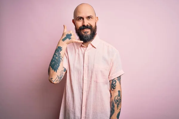 ひげと入れ墨を持つハンサムなはげ男は 孤立したピンクの背景の上にカジュアルなシャツを着て 電話で話すような手と指で電話のジェスチャーをして笑っています 概念の伝達 — ストック写真