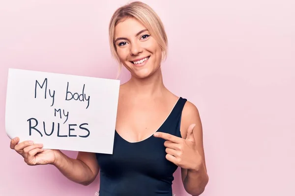 Young blonde woman asking for women freedom holding paper with my body my rules message smiling happy pointing with hand and finger