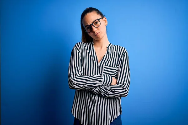 Beautiful blonde woman with blue eyes wearing striped shirt and glasses over blue background skeptic and nervous, disapproving expression on face with crossed arms. Negative person.