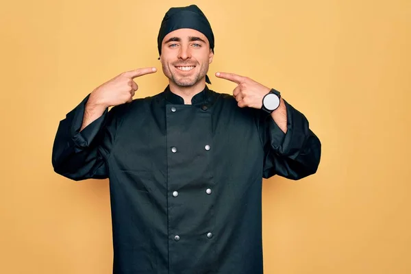 Young handsome cooker man with blue eyes wearing uniform and hat over yellow background smiling cheerful showing and pointing with fingers teeth and mouth. Dental health concept.