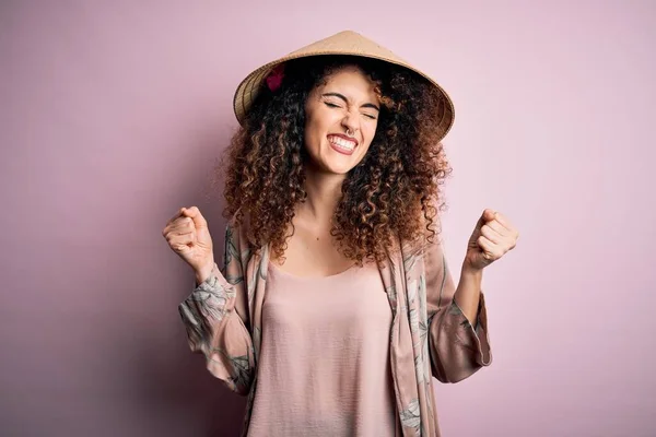 Young beautiful woman with curly hair and piercing wearing traditional asian conical hat very happy and excited doing winner gesture with arms raised, smiling and screaming for success. Celebration concept.