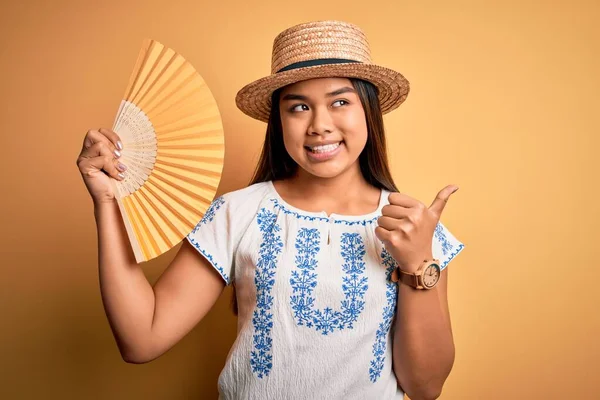 年轻的亚洲女游客在度假时戴着夏帽 手扇盖在黄色的背景上 手指朝天 笑容满面 — 图库照片