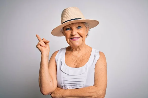 Senior beautiful grey-haired woman on vacation wearing casual summer dress and hat with a big smile on face, pointing with hand and finger to the side looking at the camera.