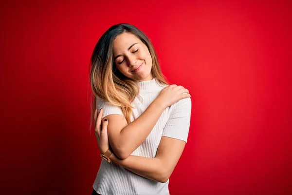 Beautiful blonde woman with blue eyes wearing casual white t-shirt over red background Hugging oneself happy and positive, smiling confident. Self love and self care