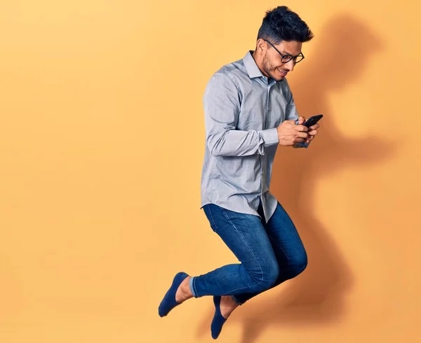 Young handsome latin man wearing glasses jumping with smile on face. Using smartphone over isolated yellow background