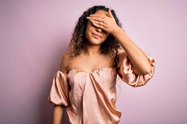 Young beautiful woman with curly hair wearing casual t-shirt standing over pink background covering eyes with hand, looking serious and sad. Sightless, hiding and rejection concept