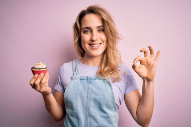 Sarışın, güzel bir kadın izole edilmiş pembe arka planda çikolatalı kek yapıyor. Tamam işareti yapıyor, mükemmel bir sembol.