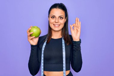 Güzel esmer sporcu kadın mezura kullanarak mor arka planda elma meyvesi yiyor. Parmaklarıyla işaret veriyor, gülümsüyor.