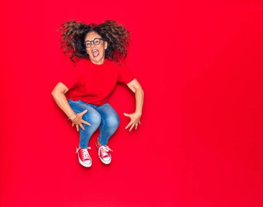 Orta yaşlı, güzel İspanyol bir kadın günlük kıyafetler ve mutlu bir gülümseme takınıyor. İzole edilmiş kırmızı arka planda yüzünde gülümsemeyle zıplıyor
