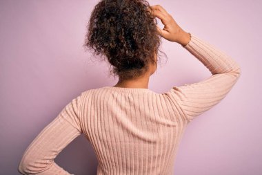 Genç, güzel, Afro-Amerikalı bir kız pembe arka planda dikiliyor ve elleri kafasında şüpheyi düşünüyor.