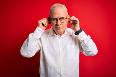 Orta yaşlı yakışıklı, beyaz gömlekli, kırmızı arka planda gözlüklü, kulaklarını parmaklarıyla çekerek gülümseyen komik bir adam. Seçme sorunu