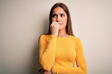 Beyaz arka plan üzerine sarı günlük tişört giyen genç, güzel esmer kadın elleri ağız yiyen tırnaklarla stresli ve gergin görünüyor. Endişe sorunu.