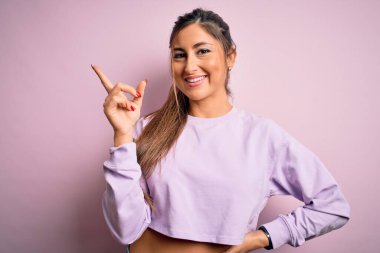 Pembe izole edilmiş arka plan üzerine kazak giyen genç ve güzel bir kadın yüzünde kocaman bir gülümseme, eli ve parmağı kameraya bakıyor..