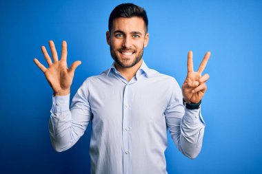 Zarif gömlekli genç adam mavi arka plan üzerinde duruyor ve yedinci parmaklarıyla işaret ederken kendinden emin ve mutlu bir şekilde gülümsüyor..