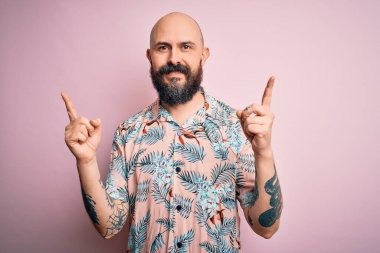 Sakallı ve dövmeli yakışıklı, pembe arka planda sıradan bir çiçek gömleği giyen, kendine güvenen, parmakları farklı yönleri gösteren kel bir adam. Reklam için boşluk kopyala