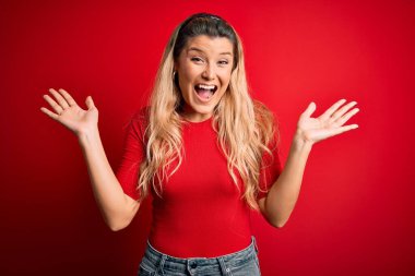 Genç ve güzel sarışın bir kadın, gündelik tişört giyip izole edilmiş kırmızı arka planda dikilmiş çılgınlığı kutluyor ve başarıya hayret ediyor. Kolları havada ve gözleri açık, heyecanlı bir şekilde çığlık atıyor. Kazanan konsept