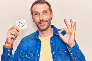 Elinde gülümseme emojisi olan genç yakışıklı adam parmaklarıyla imza atıyor, gülümseyerek mükemmel bir sembol oluşturuyor. 