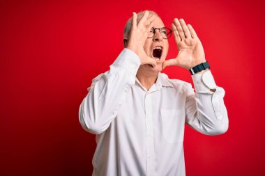 Orta yaşlı yakışıklı, beyaz gömlekli ve kırmızı arka planda gözlüklü bir adam elleri ağzında sinirli bir şekilde bağırıyor.
