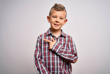 Mavi gözlü, zarif gömlekli küçük beyaz bir çocuk izole edilmiş arka planda neşeli bir şekilde duruyor. Yüzünde mutlu ve doğal bir ifadeyle eli ve parmağı yana dönük bir gülümseme var.