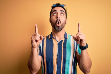 Tatilde sakallı, yaz çizgili gömlek ve güneş gözlüğü takan yakışıklı turist şaşırmış, şaşırmış ve parmaklarıyla işaret etmiş ve kollarını kaldırmış..