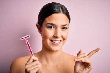 Tıraş makinesi kullanan genç ve güzel bir kız soyutlanmış pembe arka planda dururken mutlu bir şekilde elini ve parmağını kenarı işaret ediyor.