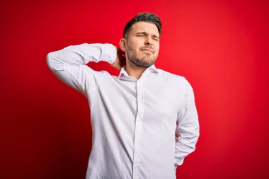 Mavi gözlü, zarif gömlekli genç bir iş adamı kırmızı izole edilmiş arka planda duruyor boynu ağrıyor, boynuna elle dokunuyor, kas ağrısı çekiyor.