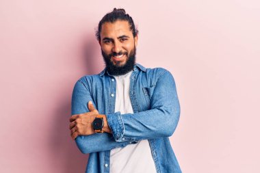 Genç Arap adam sıradan kıyafetler giyip kollarını kavuşturup kameraya bakarken gülümsüyor. pozitif kişi. 