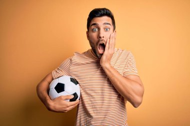 Sarı arka planda futbol topu tutan sakallı yakışıklı oyuncu. Korkmuş ve şok olmuş. Şaşırmış ve şaşırmış.