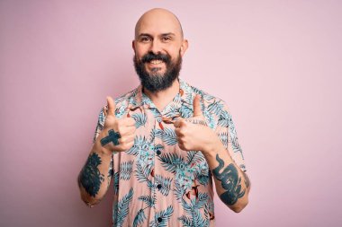 Sakallı ve dövmeli yakışıklı, pembe arka plan başarısı tabelasının üzerine gündelik çiçekli gömlek giyen, el hareketleriyle olumlu hareket eden, gülümseyen ve mutlu bir adam. Neşeli ifade ve kazanan jest.