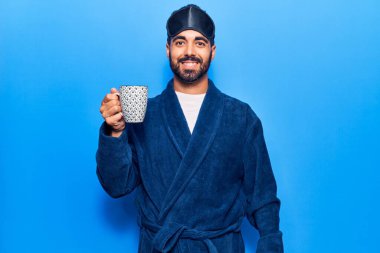 Genç İspanyol adam uyku maskesi takıyor ve sabahlık giyiyor. Kahve içiyor. Pozitif ve mutlu görünüyor. 