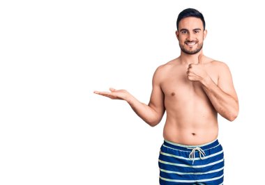 Mayo giyen genç ve yakışıklı bir adam avuç içini gösteriyor ve baş parmaklarıyla iyi bir hareket yapıyor, mutlu ve neşeli gülümsüyor. 