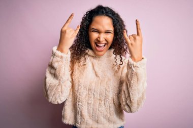 Kıvırcık saçlı genç bir kadın pembe arka planda dikilip elleri havada rock sembolü yaparak bağırıyor. Müzik yıldızı. Ağır müzik konsepti.