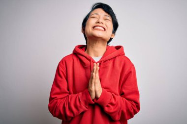 Genç, güzel Asyalı bir kız beyaz arka planda kapüşonlu bir kazak giyiyor. Elleri bağlı yalvarıyor ve dua ediyor. Yüzünde umut ifadesi var. Yalvarıyorum..