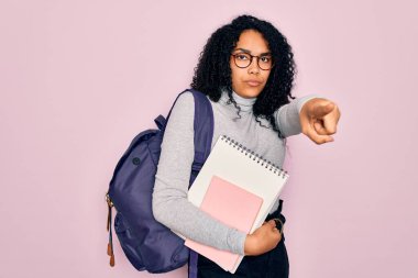 Sırt çantalı ve gözlüklü genç Afro-Amerikan kıvırcık saçlı kadın kamerayı işaret eden bir kitap tutuyor ve sen, el işareti, ön taraftan pozitif ve kendinden emin bir jest.