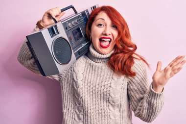 Güzel kızıl saçlı bir kadın pembe arka planda klasik müzik dinlerken mutlu bir gülümseme ve kaldırılmış bir el ile kazanan bir ifadeyle başarıyı kutluyor.