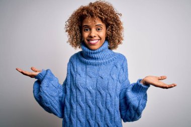 Güzel Afro-Amerikan kadın balıkçı yaka kazak giyiyor beyaz arka planda gülümsüyor iki elini de açık avuçlarında gösteriyor, sunuyor ve reklam karşılaştırması yapıyor ve dengeyi sağlıyor.