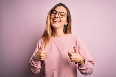 Mavi gözlü güzel sarışın kadın pembe arka plan başarı tabelasına gözlük ve süveter takıyor. Elleriyle olumlu jest yapıyor. Gülümsüyor ve mutlu. Neşeli ifade ve kazanan jest.