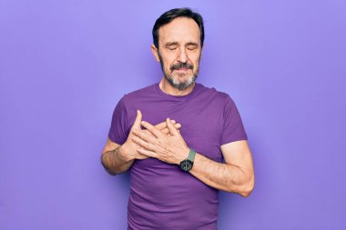 Orta yaşlı, mor tişört giyen yakışıklı bir adam izole edilmiş arka planda duruyor. Elleri göğsünde gülümsüyor, yüzünde minnet dolu bir jest ile gözleri kapalı. Sağlık kavramı.